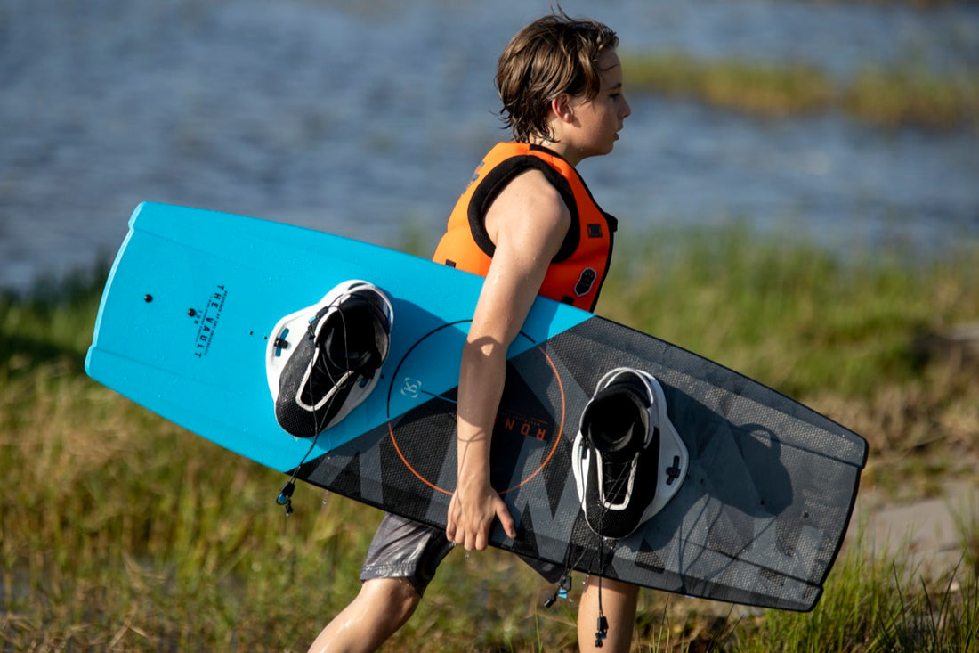 Image of a kid in orange lifejacket carrying a blue and black wakeboard