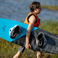 Image of a kid in orange lifejacket carrying a blue and black wakeboard